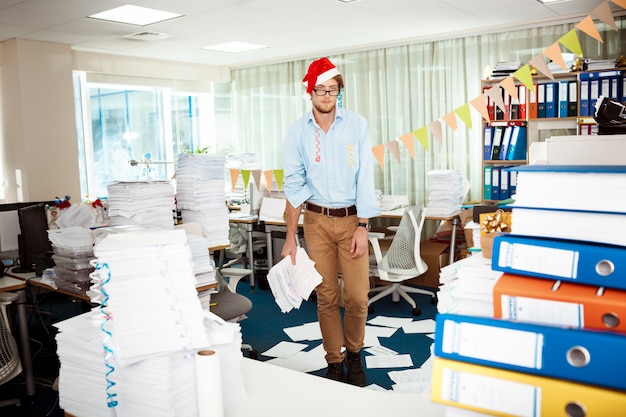 Hombre de negocios joven cansado que trabaja en oficina entre los papeles el día de Navidad.