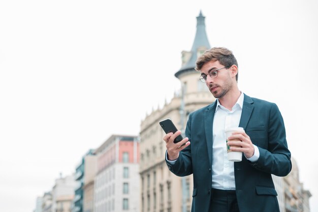 Hombre de negocios joven chocado que mira el smartphone que sostiene la taza de café disponible