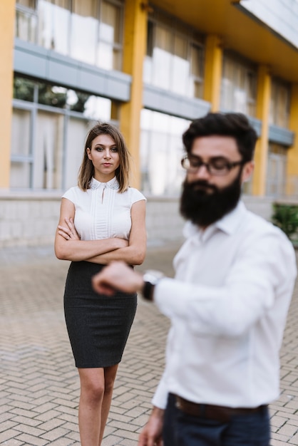 Foto gratuita hombre de negocios joven defocused que mira el reloj con la mujer joven elegante que se coloca en el fondo