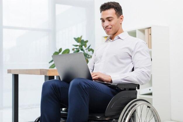 Hombre de negocios joven discapacitado feliz que se sienta en la silla de ruedas usando el ordenador portátil en la oficina