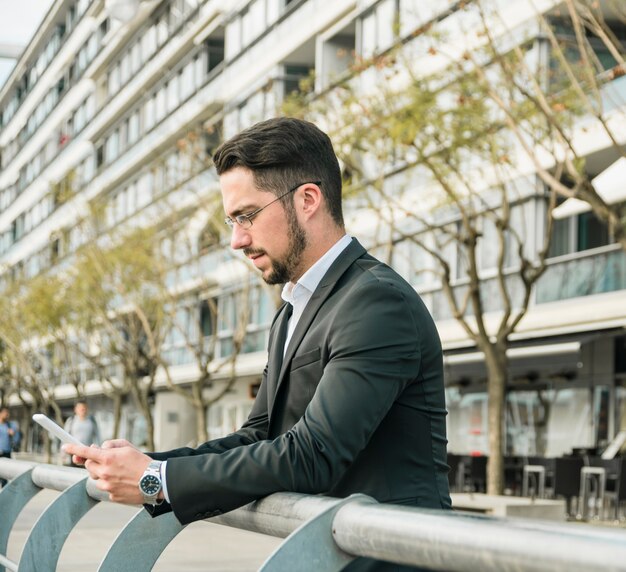 Hombre de negocios joven elegante que se coloca cerca de la verja usando el teléfono móvil