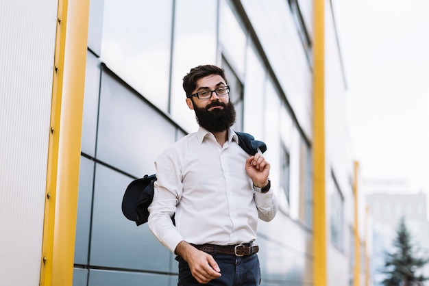 Foto gratuita hombre de negocios joven elegante que sostiene la capa sobre su hombro que se coloca delante del edificio