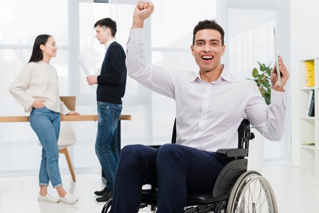 Hombre de negocios joven emocionado que sostiene la tableta digital en la mano que se sienta en la silla de ruedas con los pares del negocio que miran uno a