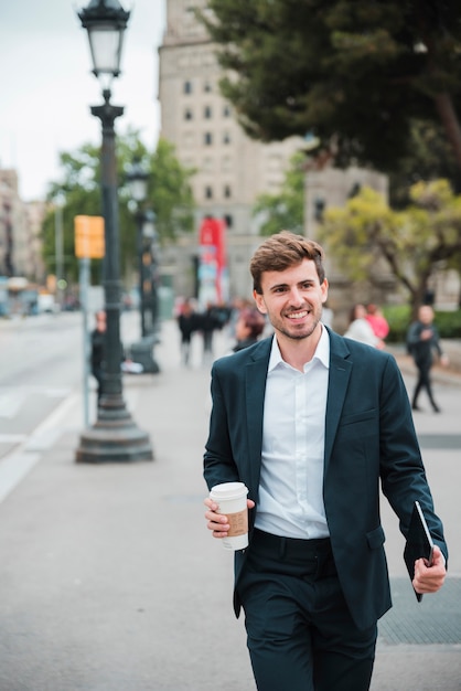 Hombre de negocios joven feliz que camina en la calle con su tableta digital y la taza de café disponible