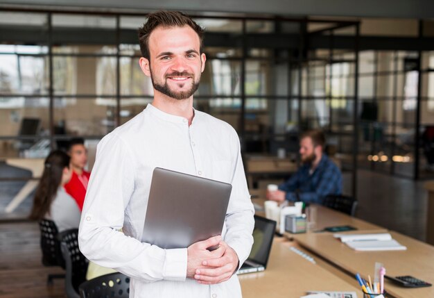 Hombre de negocios joven feliz que mira la cámara que sostiene el ordenador portátil en oficina