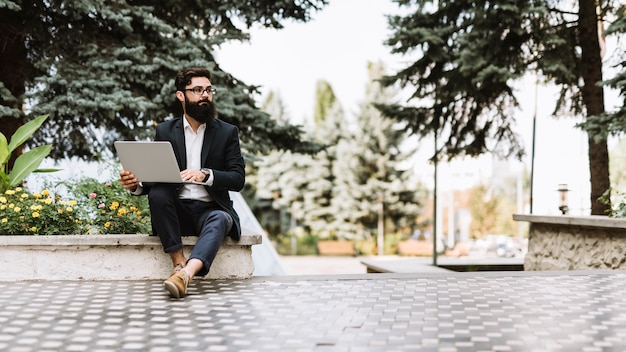 Hombre de negocios joven hermoso que se sienta con el ordenador portátil en el parque que mira lejos
