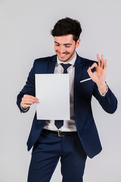 Hombre de negocios joven hermoso que sostiene el Libro Blanco en la mano que muestra la muestra aceptable contra el contexto gris