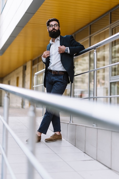 Hombre de negocios joven moderno con la barba larga que se coloca delante del edificio corporativo