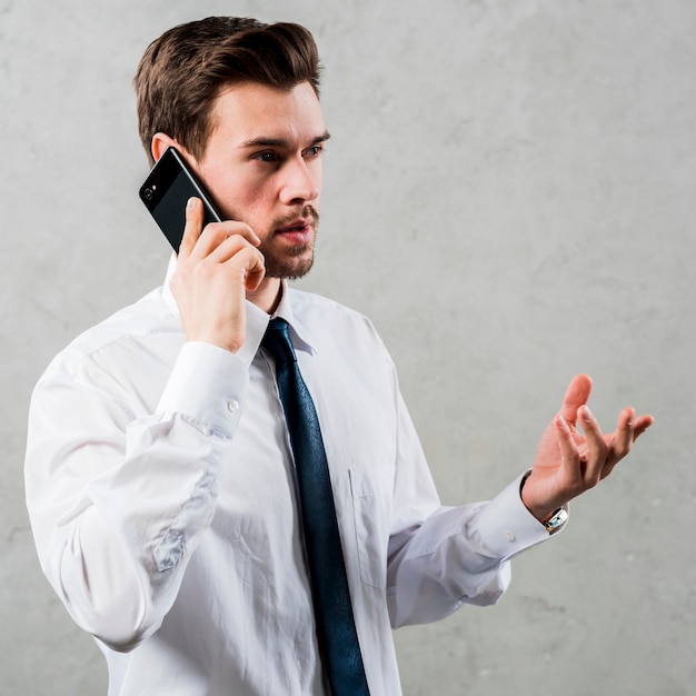 Hombre de negocios joven que habla en el teléfono elegante que gesticula la situación contra la pared gris