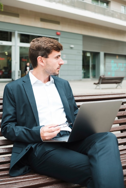 Hombre de negocios joven que se sienta en banco con la computadora portátil