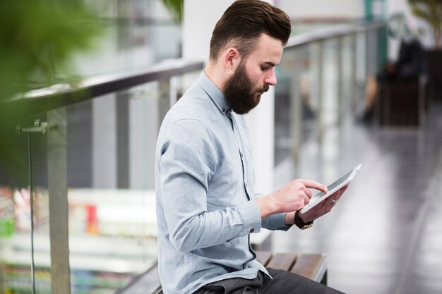 Hombre de negocios joven que se sienta en banco en el pasillo usando la tableta digital