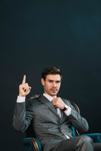 Hombre de negocios joven que se sienta en la butaca que muestra la mano con un finger para arriba