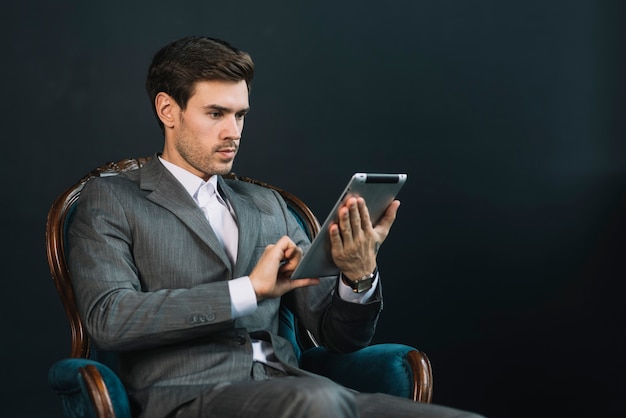 Hombre de negocios joven que se sienta en la butaca que usa la tableta digital contra fondo oscuro