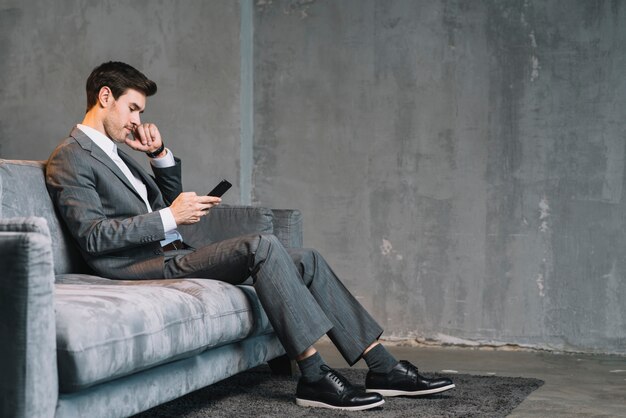 Hombre de negocios joven que se sienta en el sofá gris usando el teléfono móvil