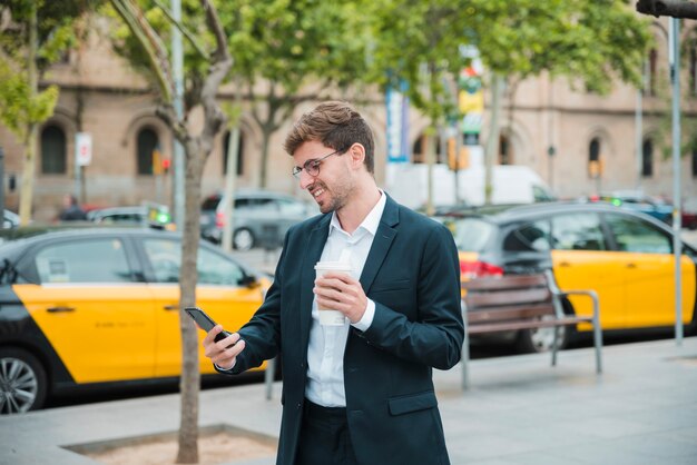 Hombre de negocios joven que sostiene la taza de café que mira el teléfono móvil