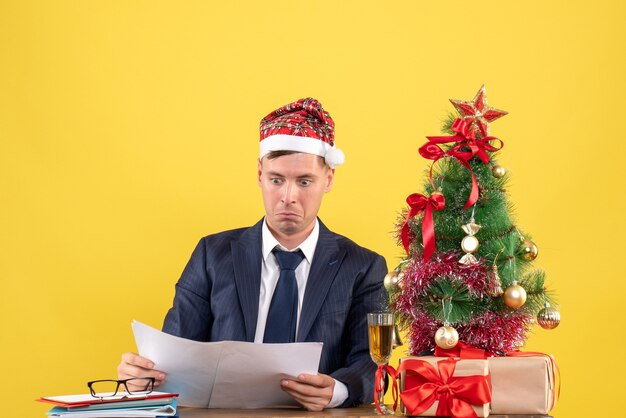 Hombre de negocios joven revisando los papeles sentado en la mesa cerca del árbol de Navidad y presenta en amarillo