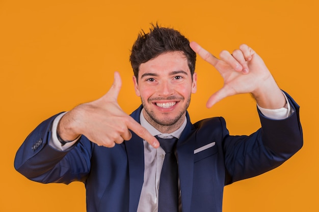 Hombre de negocios joven sonriente confiado que hace el marco de la mano contra un contexto anaranjado