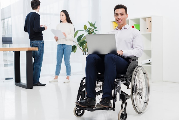 Un hombre de negocios joven sonriente discapacitado que se sienta en la silla de ruedas con el ordenador portátil delante del colega del negocio