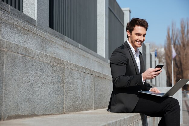 Hombre de negocios joven sonriente que se sienta al aire libre charlando por teléfono.