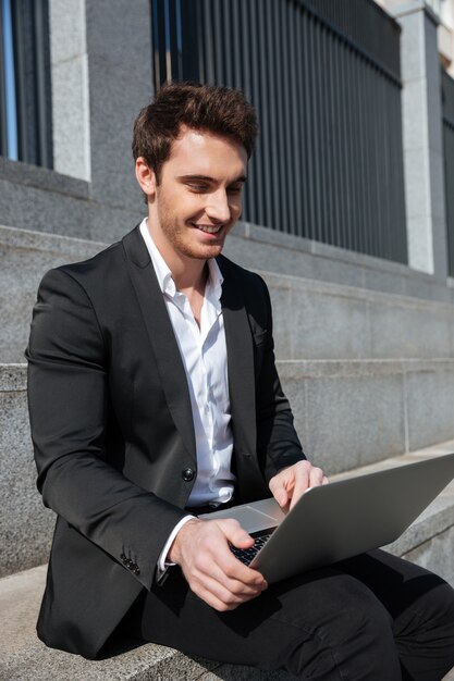 Hombre de negocios joven sonriente que se sienta al aire libre usando la computadora portátil