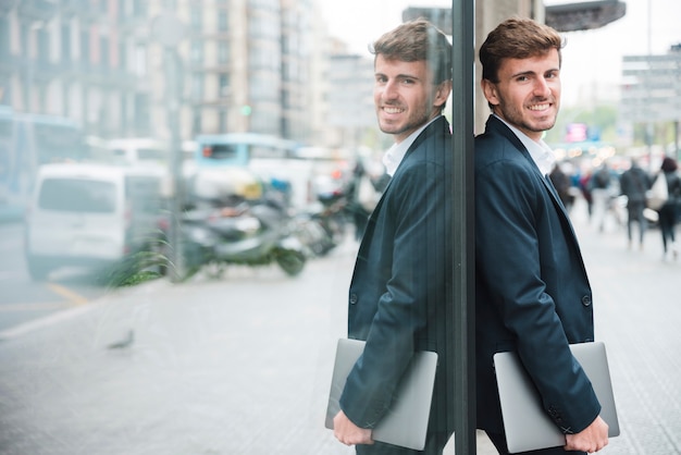 Hombre de negocios joven sonriente que sostiene el teléfono móvil en la mano que se inclina en la pared de cristal