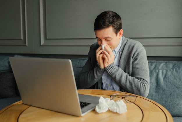 El hombre de negocios joven sopla su nariz mientras que trabaja en su computadora portátil en el lugar de trabajo.