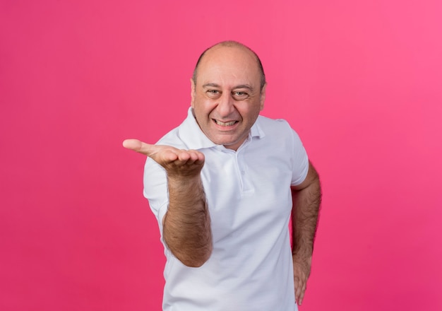 Hombre de negocios maduro casual molesto manteniendo la mano en la cintura y mostrando la mano vacía en la cámara aislada sobre fondo rosa con espacio de copia