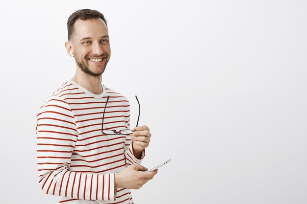 Hombre de negocios masculino exitoso positivo, quitándose las gafas, mirando a un lado y sonriendo amigablemente, eligiendo música para escuchar en el teléfono inteligente, usando auriculares inalámbricos