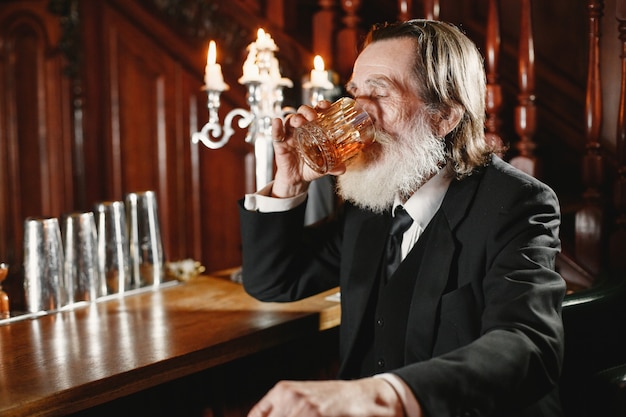 Hombre de negocios mayor barbudo. El hombre bebe un whisky. Senior en traje negro.