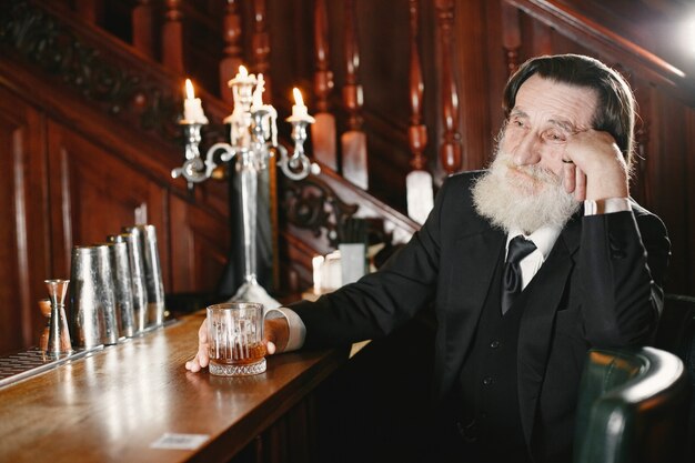 Hombre de negocios mayor barbudo. El hombre bebe un whisky. Senior en traje negro.