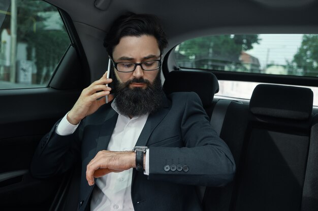 Un hombre de negocios mientras viaja en automóvil en el asiento trasero con un teléfono inteligente