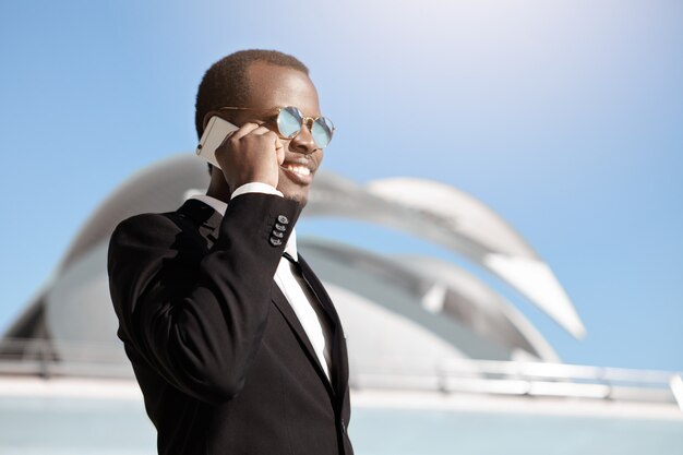 Hombre de negocios negro alegre feliz en ropa formal y gafas de sol hablando por teléfono inteligente fuera de la oficina que construye temprano en la mañana, haciendo una cita para una reunión de negocios con socios potenciales
