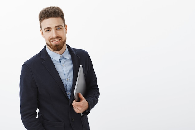 Hombre de negocios con ojos azules y barba de pie seguro de sí mismo en traje formal sosteniendo un portátil en la mano mirando complacido y seguro, siendo ambicioso y exitoso