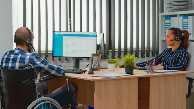 Foto gratuita hombre de negocios paralizado en silla de ruedas con auricular que realiza una cita a distancia y ofrece atención al cliente. freelancer discapacitado inmovilizado que trabaja en un edificio corporativo financiero utilizando tecnología moderna