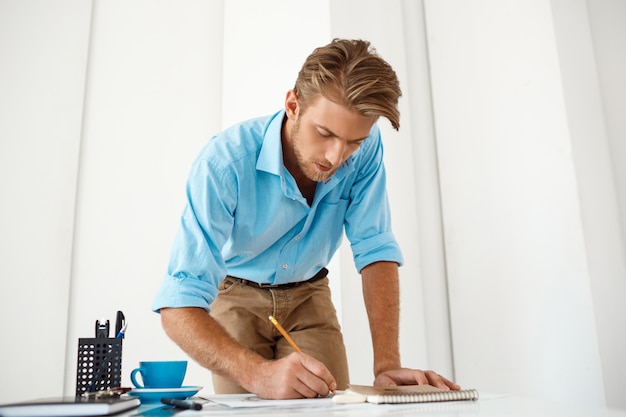 Foto gratuita hombre de negocios pensativo confiado hermoso joven que trabaja de pie en la escritura de la tabla en la libreta. interior de oficina moderno blanco.