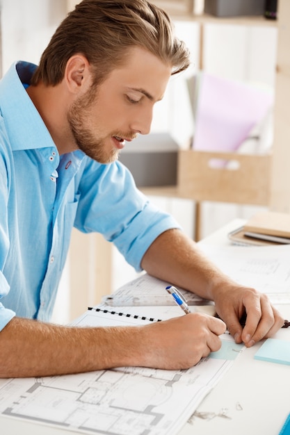 Foto gratuita hombre de negocios pensativo confidente hermoso joven que trabaja sentado en la escritura de la tabla en el cuaderno. interior de oficina moderno blanco