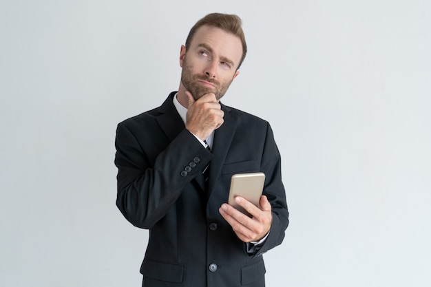 Hombre de negocios pensativo que toca la barbilla, pensando y sosteniendo smartphone.