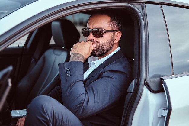 Hombre de negocios pensativo con tatuaje en el brazo con gafas de sol vestido con un traje formal sentado en los asientos delanteros del camión de lujo.