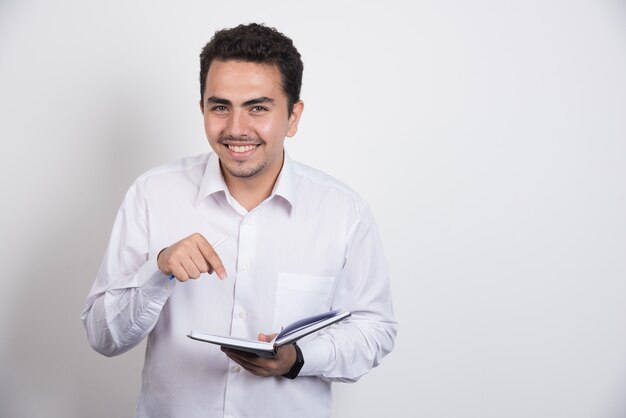 Hombre de negocios positivo apuntando al cuaderno sobre fondo blanco.