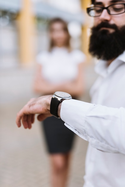 Un hombre de negocios que comprueba tiempo en el reloj con el colega borroso en el fondo