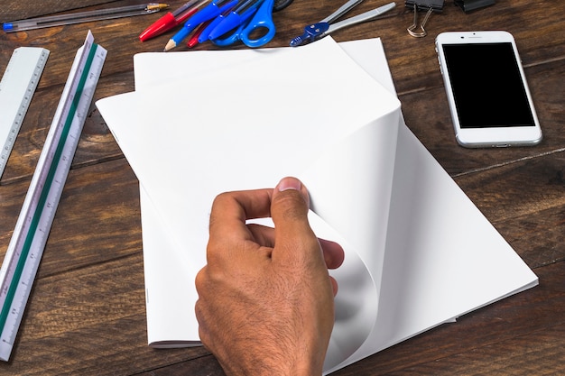 Hombre de negocios que da vuelta al papel en blanco blanco con los stationeries y el smartphone en la tabla de madera