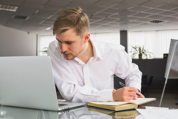 Hombre de negocios que mira la computadora portátil mientras que escribe horario en diario