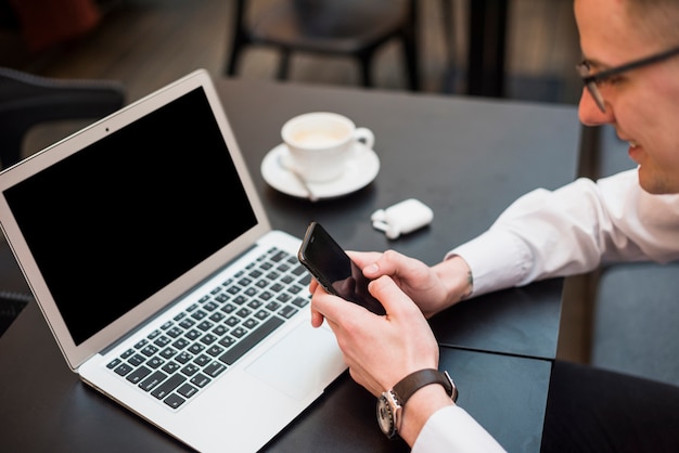 Un hombre de negocios que usa el teléfono móvil frente a la computadora portátil con una taza de café en la mesa