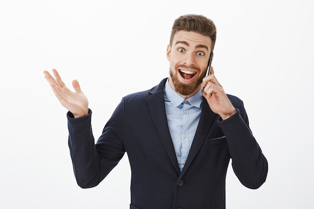 Hombre de negocios recibiendo excelentes noticias. Feliz y emocionado emprendedor guapo encantado en elegante traje sosteniendo teléfono inteligente cerca de la oreja hablando sintiéndose optimista levantando la mano y sonriendo de alegría