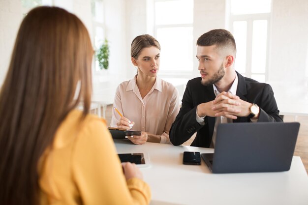 Hombre de negocios reflexivo con computadora portátil y mujer de negocios con lápiz y carpeta en las manos consultando cuidadosamente sobre el solicitante Jóvenes empleadores que pasan una entrevista de trabajo en una oficina moderna