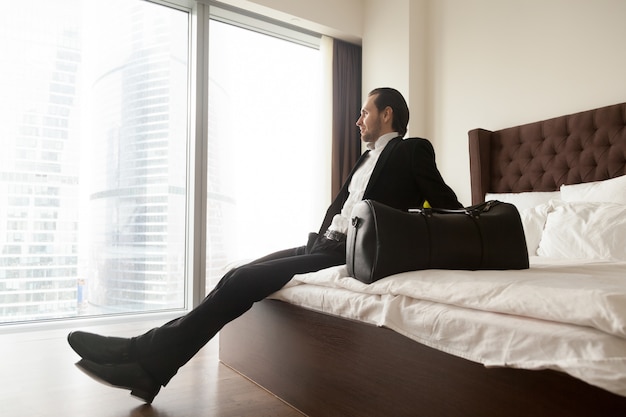 Hombre de negocios relajado que se sienta en cama además del bolso del equipaje.