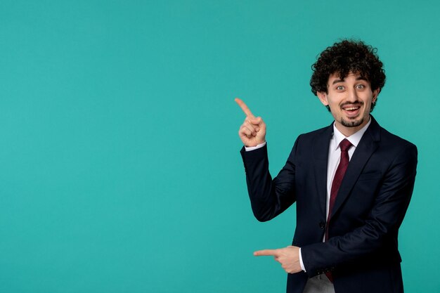 Hombre de negocios rizado lindo chico guapo en traje negro y corbata roja apuntando hacia arriba y sonriendo