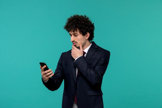 Hombre de negocios rizado lindo chico guapo en traje negro y corbata roja mirando seriamente la pantalla del teléfono