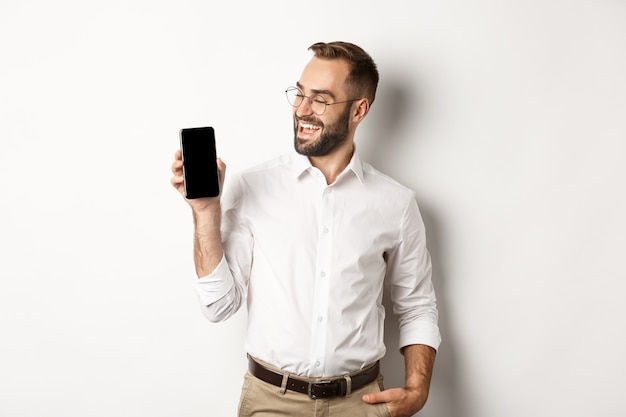 Hombre de negocios satisfecho que muestra y mira la pantalla del móvil, introduce la promoción de la aplicación o el sitio web, de pie sobre un fondo blanco.