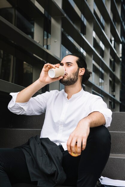 Hombre de negocios sentado cerca del edificio de oficinas con manzana en la mano tomando café